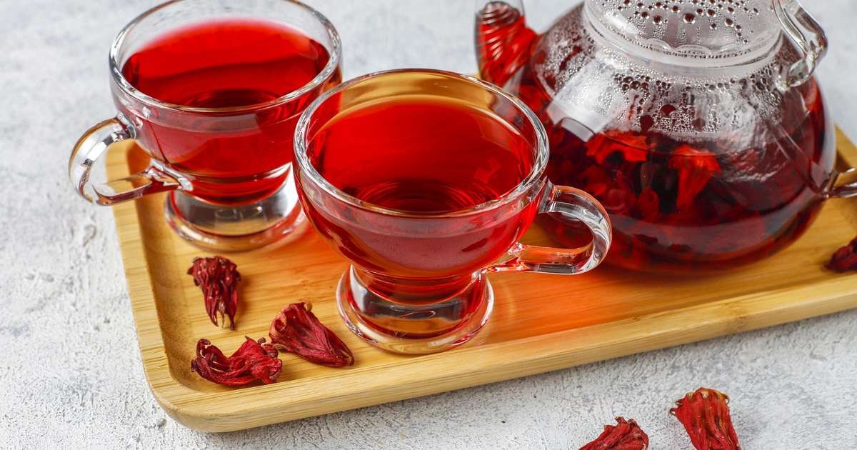 Xícara de chá de hibisco cercada por flores secas da planta Hibiscus sabdariffa em uma mesa de madeira